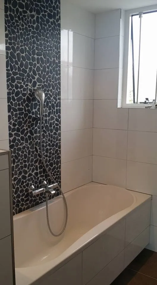 De mozaïekstrook loopt over de volle hoogte door achter het bad; de douchekraan en handdouche zijn erop uitgelijnd.