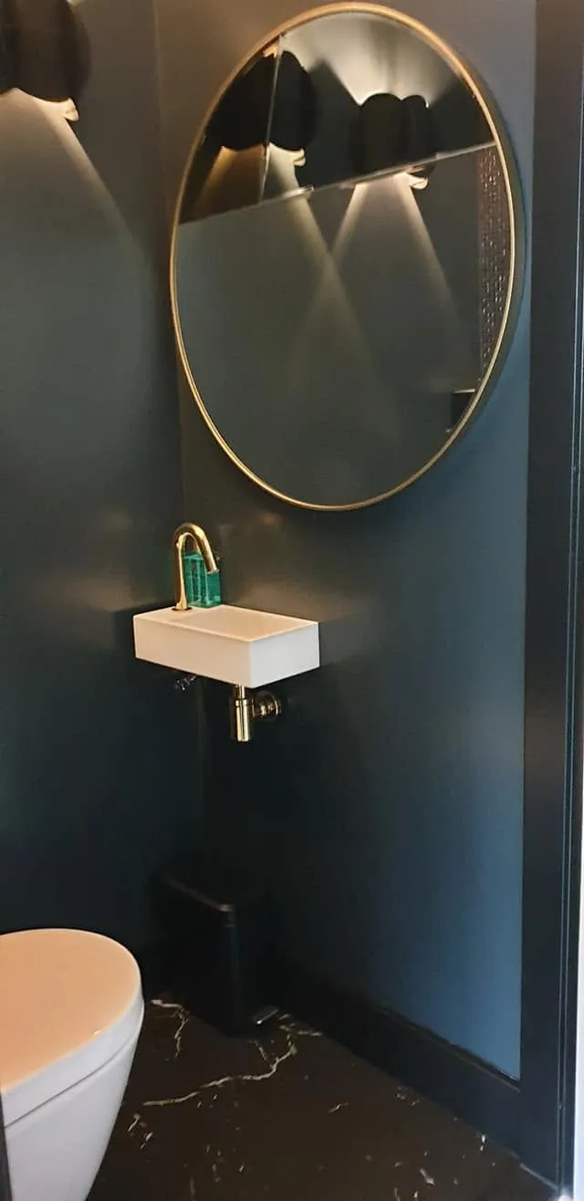 Een klein wandfonteintje met messingkleurige kraan en sifon, onder een ronde spiegel met messingkleurige rand.
