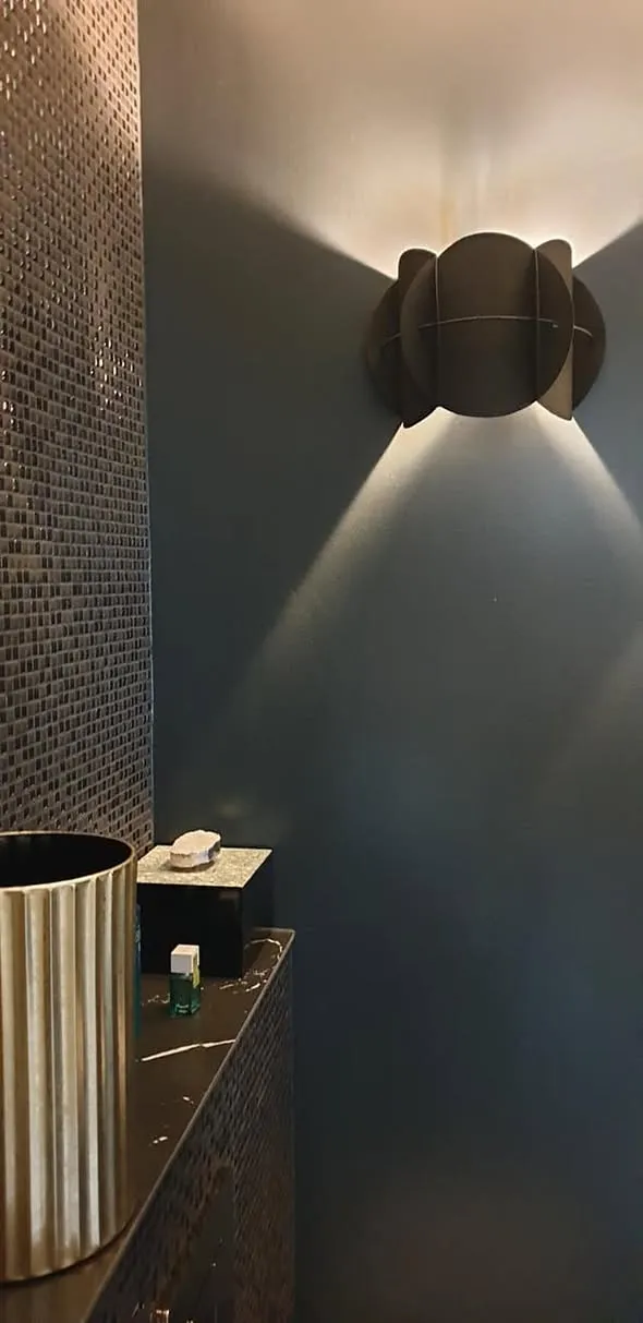 De wandlamp werpt licht omhoog en omlaag over de donkere wand; links loopt de mozaïekstrook door tot aan het plafond.