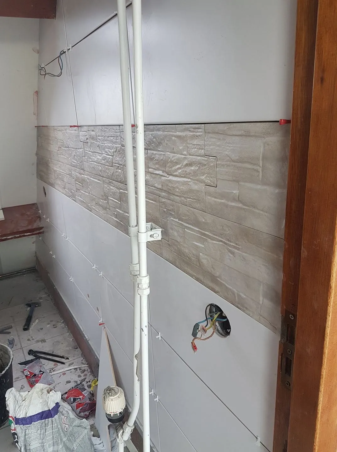 De wand wordt betegeld; de decoratieve band met reliëf zit op hoogte en de leidingen lopen nog zichtbaar over het tegelwerk.
