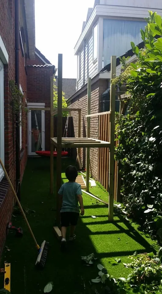 Het houten frame van het speeltoestel staat op de nieuwe grasmat; de staanders zijn gesteld en de eerste liggers zitten erin.