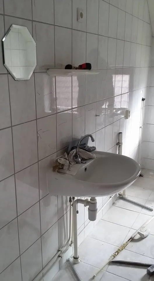 De oude badkamer voor aanvang: kleine witte wandtegels over de volle hoogte en een hangende wastafel met open leidingwerk eronder.