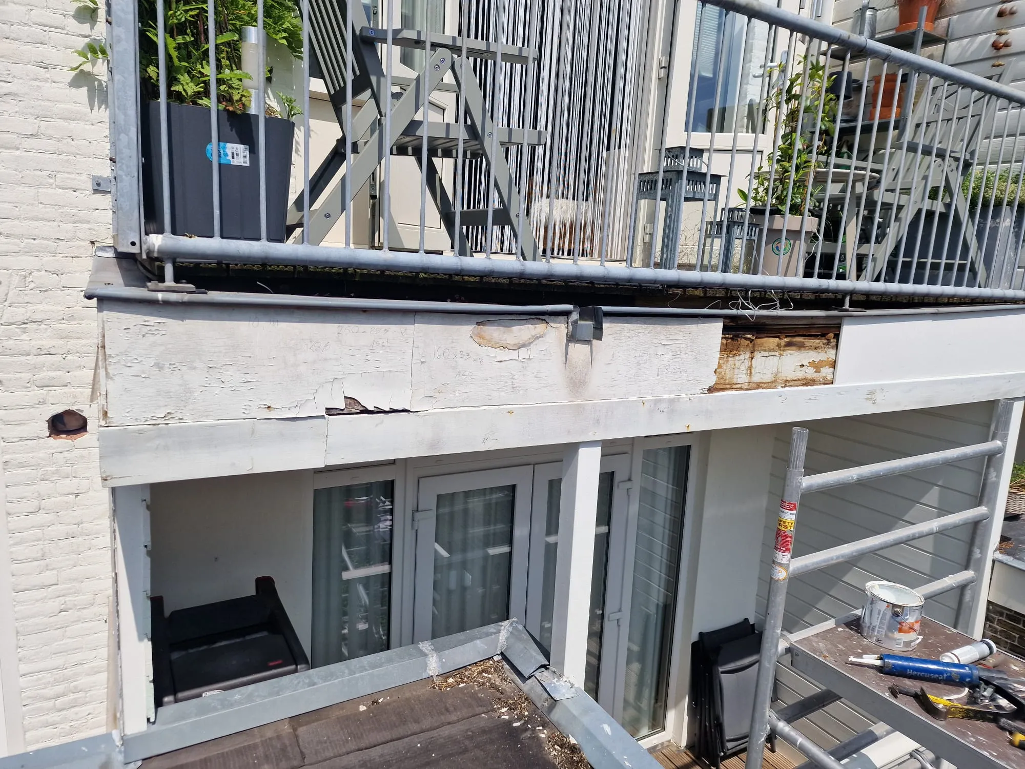 De boeiboord onder het balkon voor aanvang: de verflaag is opengebarsten en op meerdere plekken ligt het hout eronder open.