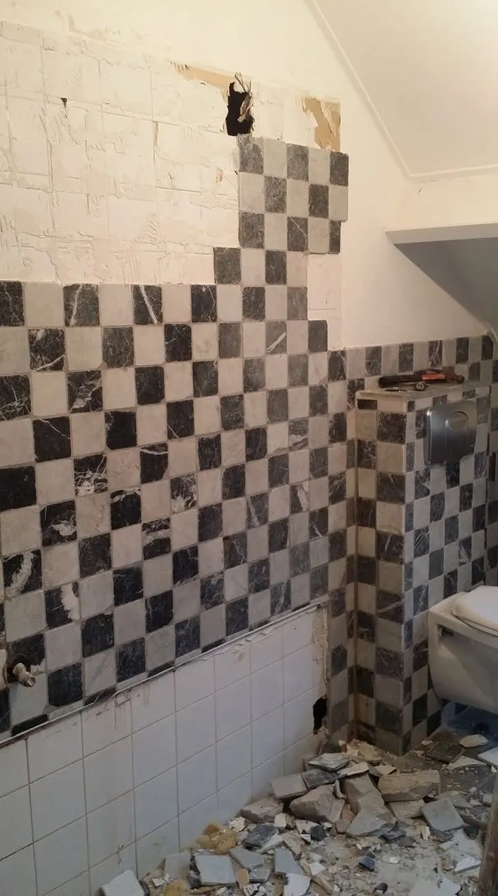 Het oude tegelwerk in zwart-wit dambordpatroon wordt van de wand gehaald; de scherven liggen op de vloer.